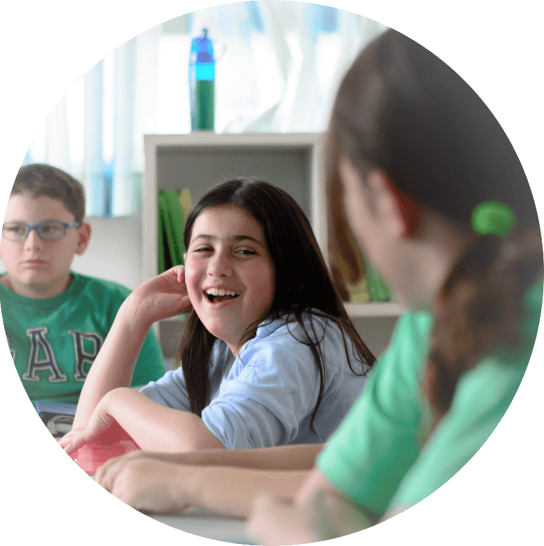Two students laughing together across a classroom table while a third works beside them